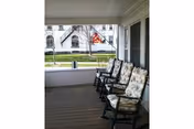 A covered porch area with four cushioned rocking chairs lined up facing outward. The porch has wooden flooring and white railings. In the background, there is a view of a street with a building and some trees.