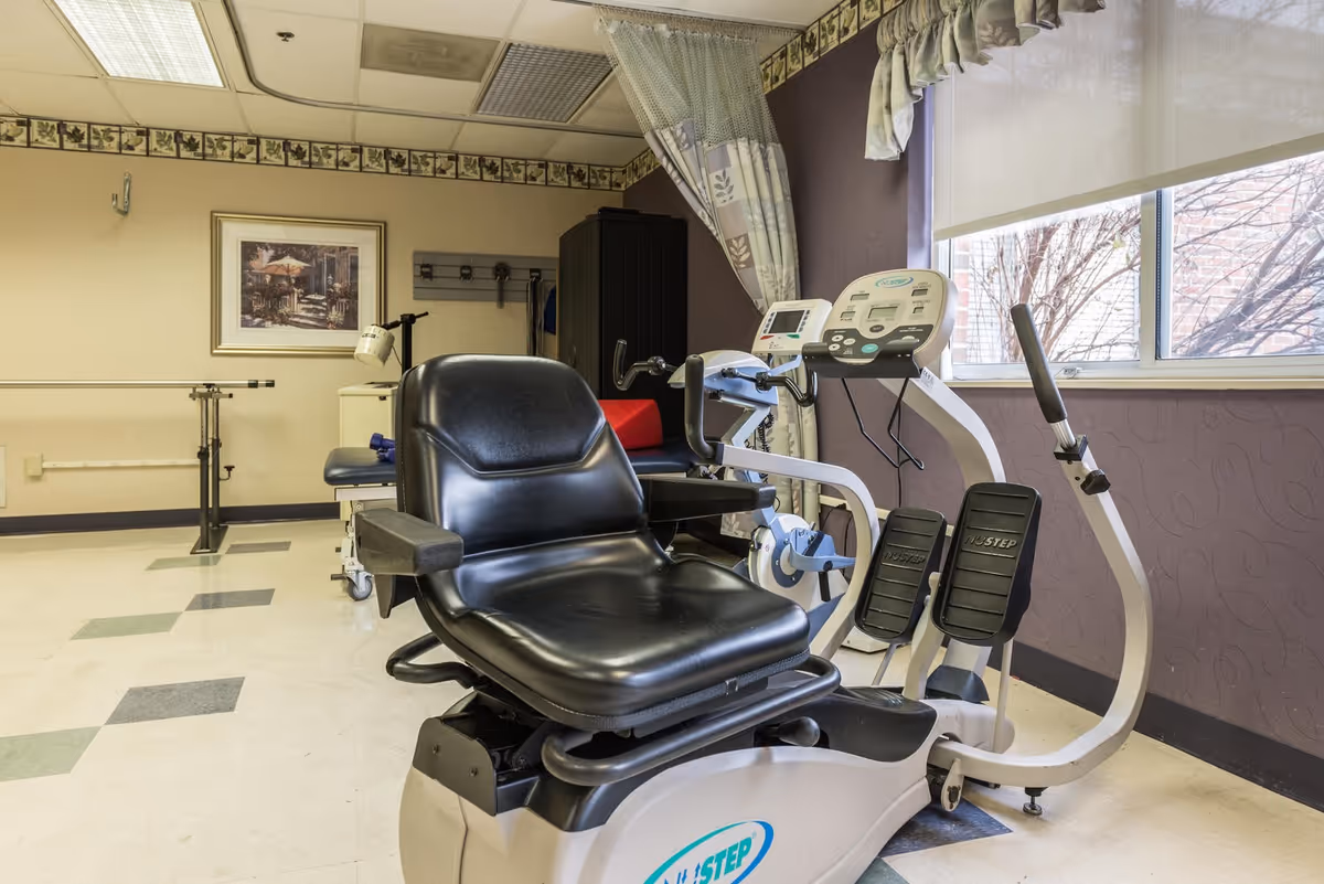 A senior living facility therapy room with a recumbent exercise machine and other rehabilitation equipment by a window.