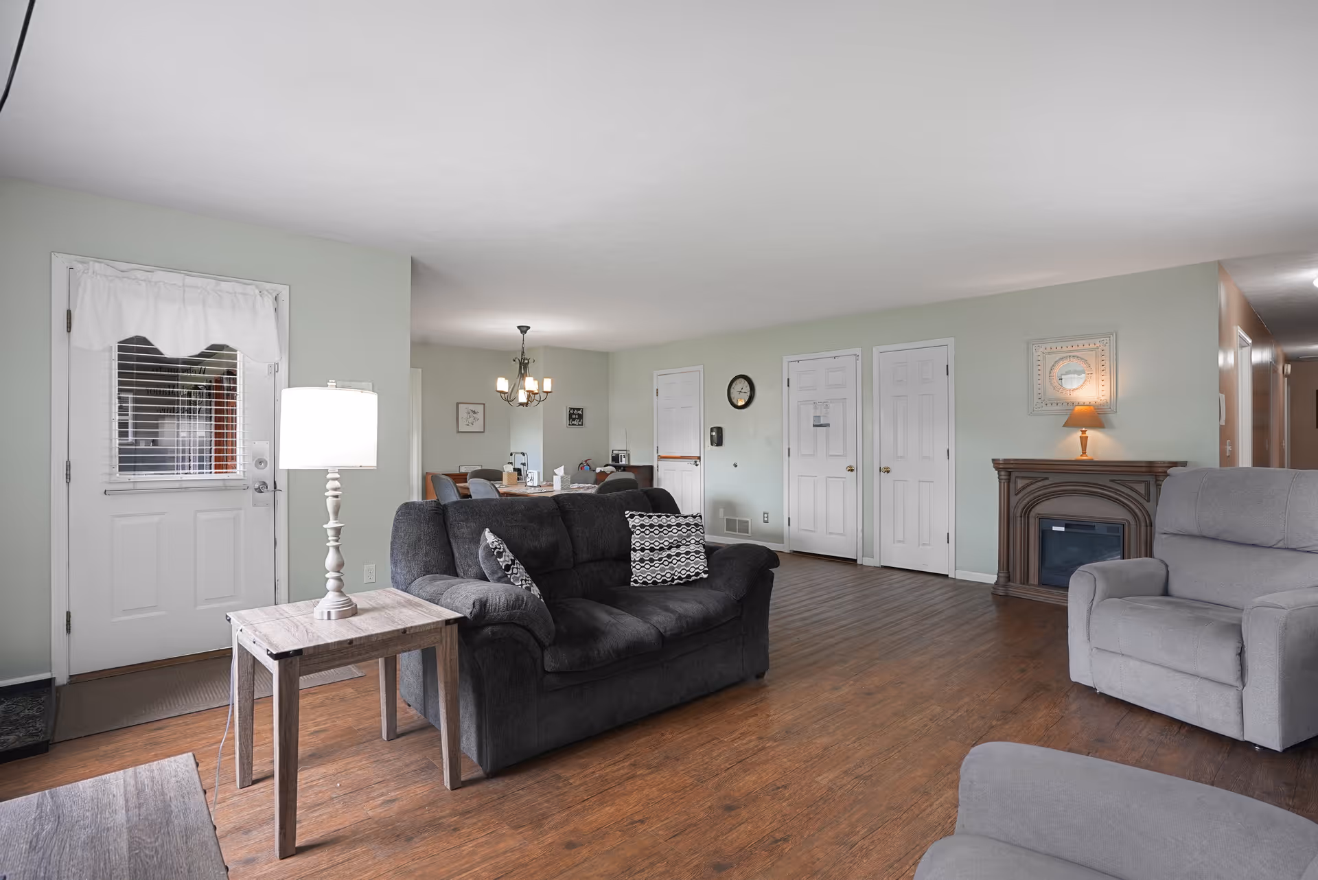 A spacious living room with wooden flooring, featuring a dark gray sofa with patterned cushions, a light gray armchair, a wooden side table with a white lamp, and a decorative fireplace with a lamp and framed mirror above it. In the background, there is a dining area with a chandelier and a table with chairs. The walls are painted light green, and there is a white door with a window and curtain on the left side.
