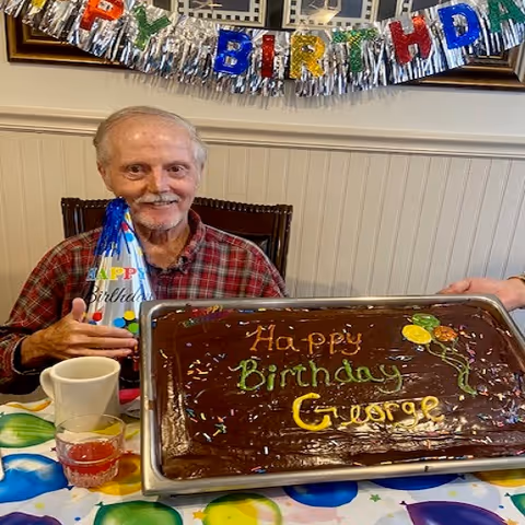 An elderly man wearing a red plaid shirt and a colorful birthday hat sits at a table with a large rectangular chocolate birthday cake in front of him. The cake has colorful icing that reads 'Happy Birthday George' and is decorated with balloons and sprinkles. Behind him, a 'Happy Birthday' banner is hanging on the wall.