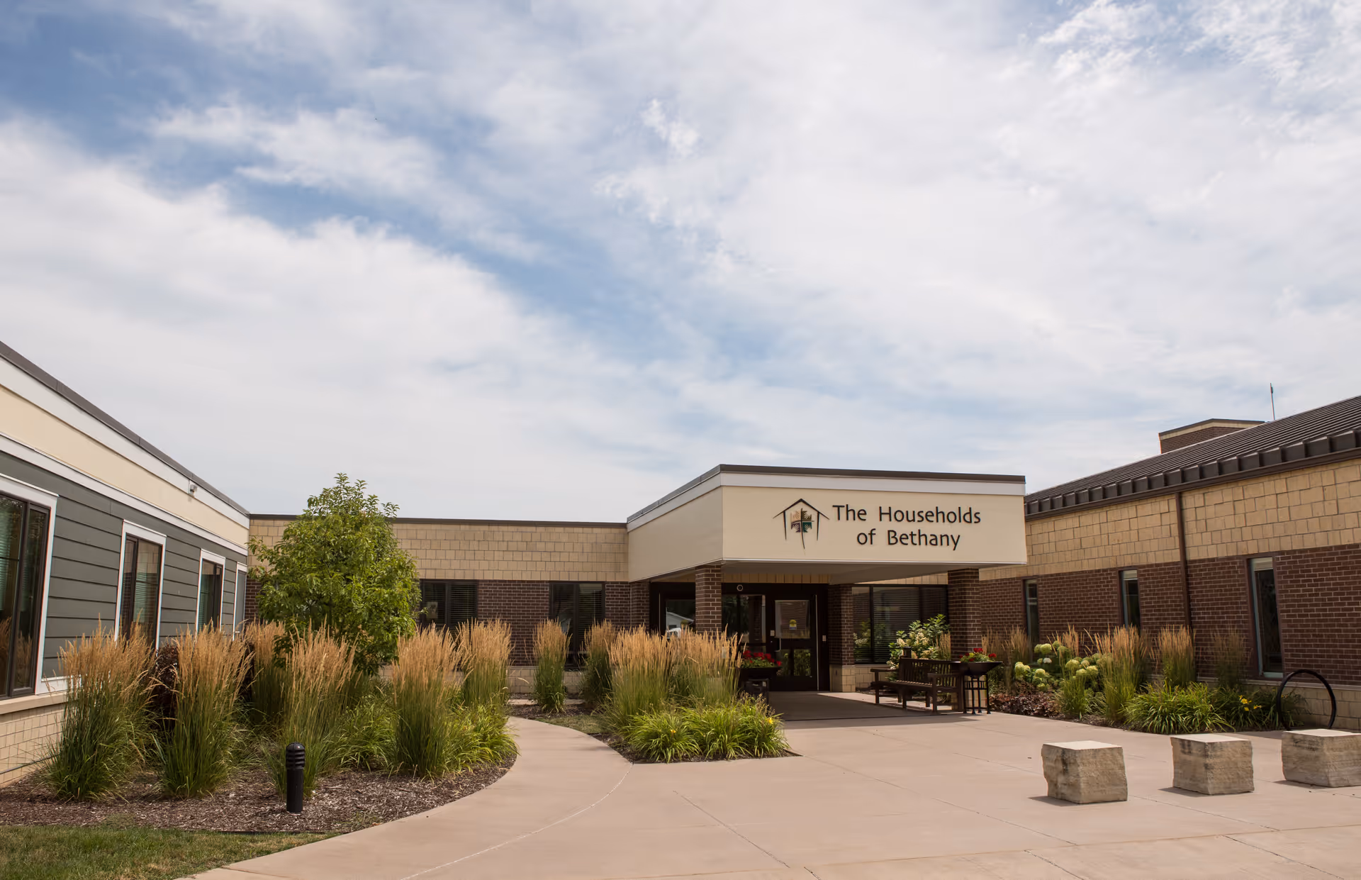 Front entrance courtyard of The Households of Bethany senior living building with landscaping and a covered entry.