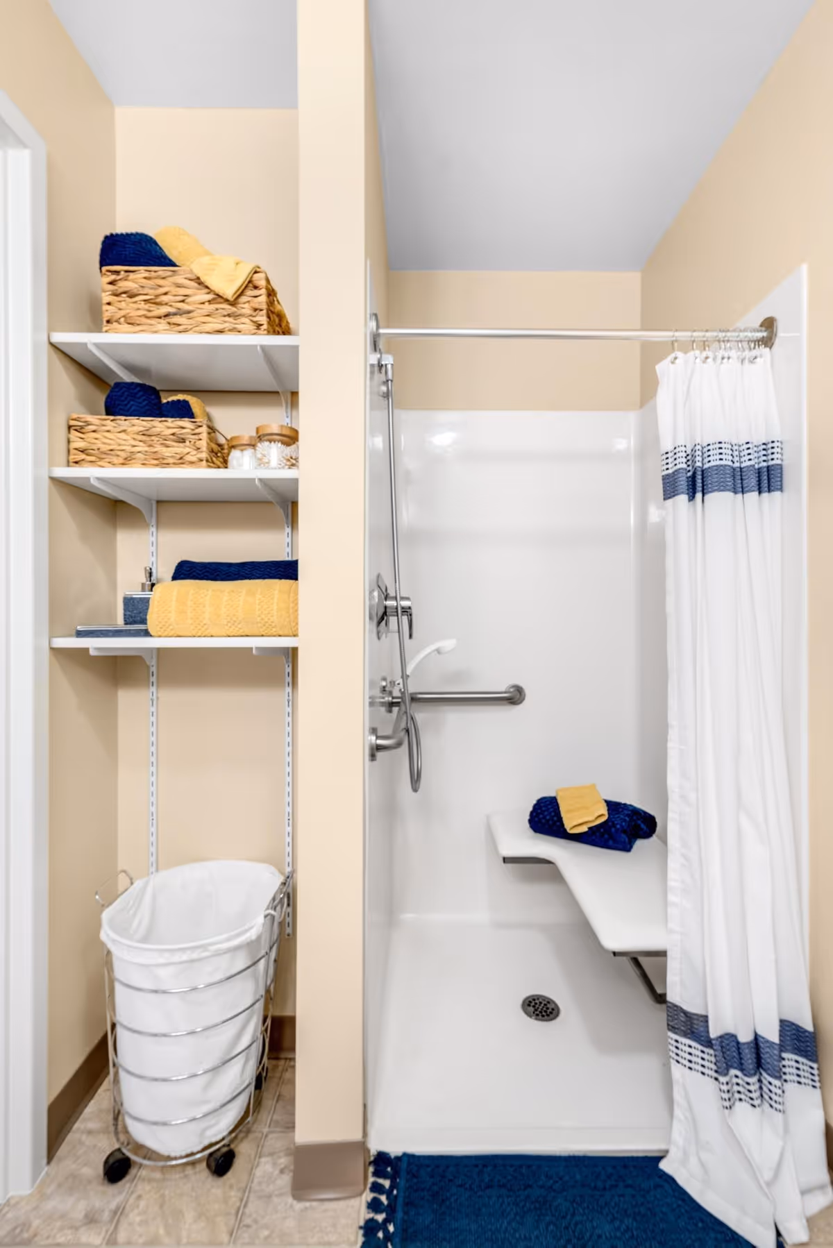 A bathroom shower area with a white shower curtain featuring blue stripes, a built-in white shower bench with folded blue and yellow towels on it, and a silver grab bar. To the left, there are white shelves holding woven baskets with towels and jars, and a white laundry hamper with a metal frame on wheels below the shelves. The walls are beige and the floor has light-colored tiles with a blue bath mat in front of the shower.
