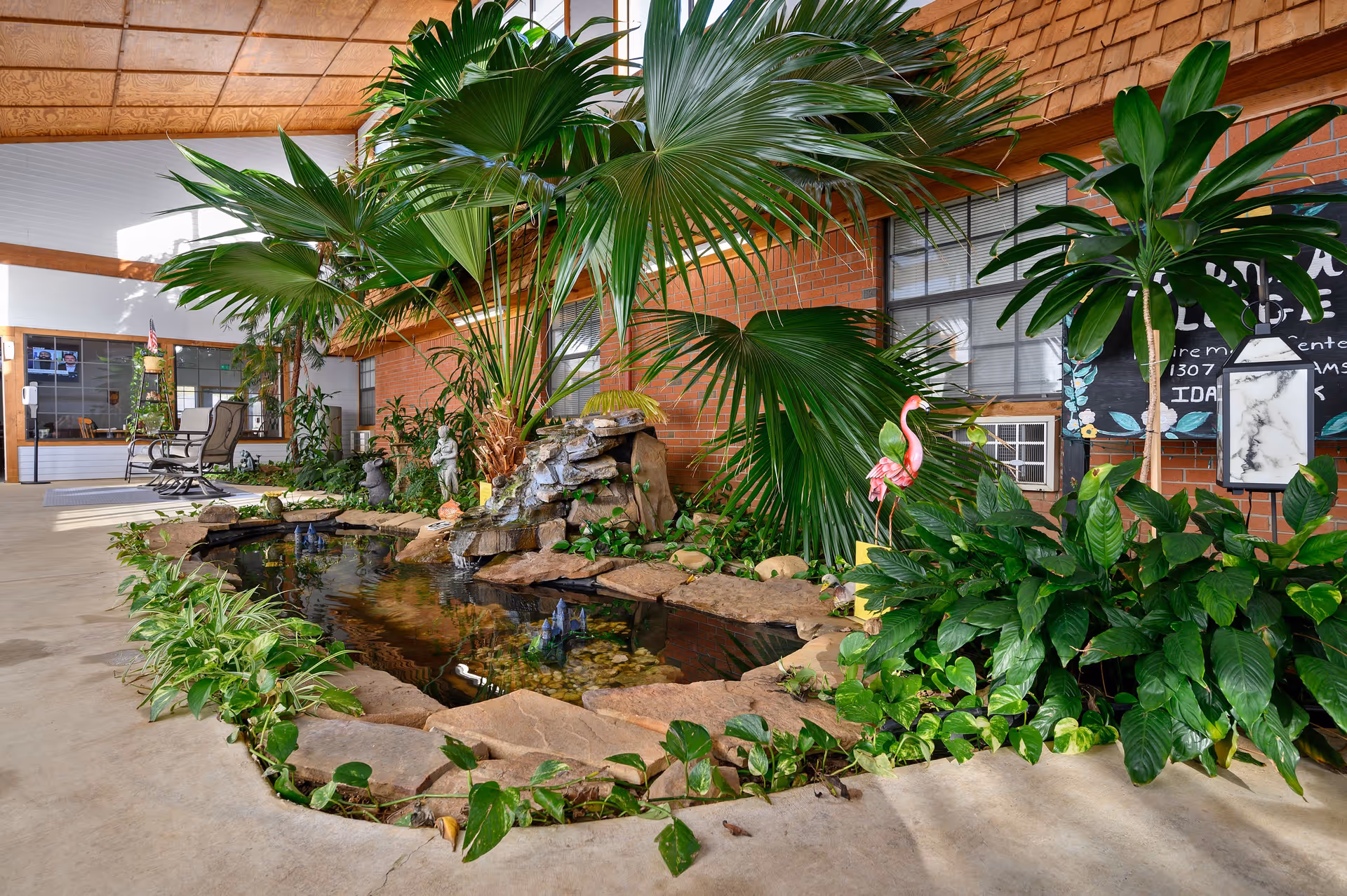 Indoor garden area with a small pond surrounded by rocks and lush green plants, including large palm leaves. There is a pink flamingo lawn ornament and a small waterfall feature. In the background, there are chairs, a window, and a chalkboard sign with partially visible text. The ceiling has a wooden panel design.