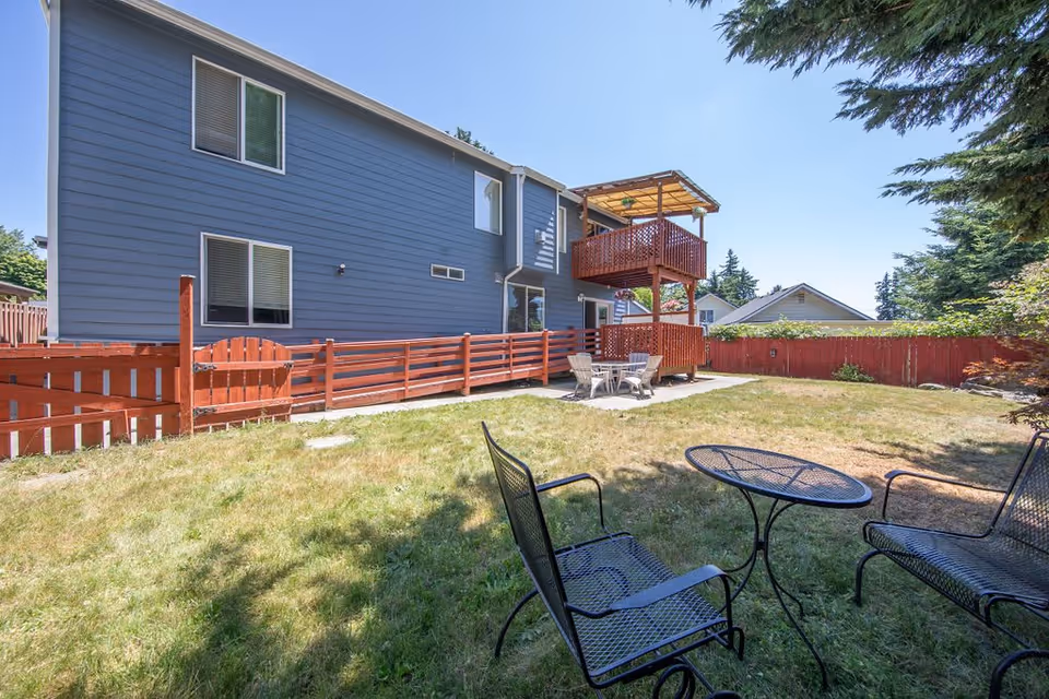 Outdoor view of a senior living facility yard with a blue two-story building featuring a red wooden balcony and fence. The yard has a grassy area with a black metal table and chairs set under the shade of trees, and a concrete patio with a white table and chairs set near the building.