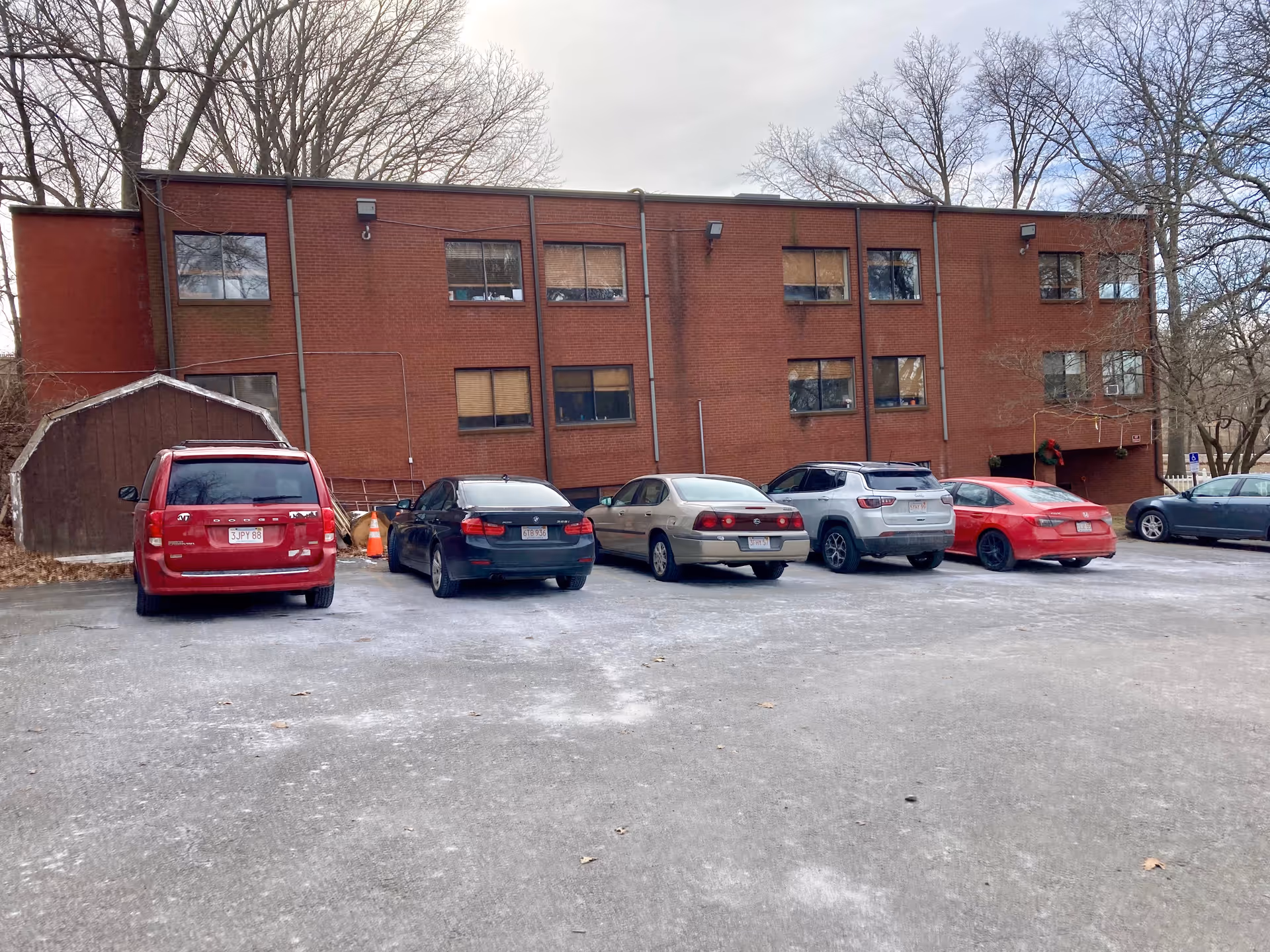 Parking lot with six parked cars in front of a two-story red brick building with multiple windows and leafless trees in the background under a cloudy sky.