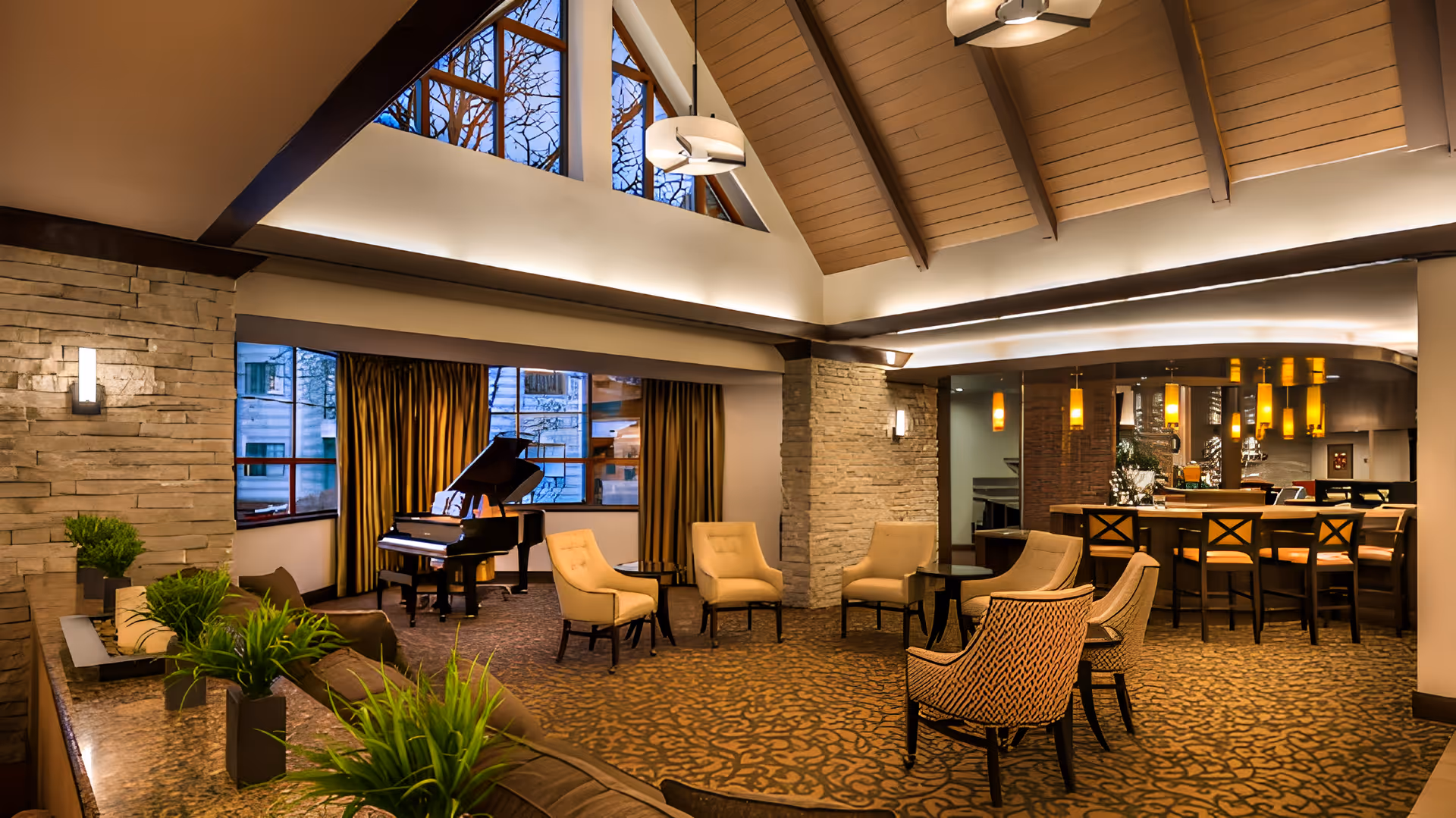 A spacious and warmly lit living room area with a high vaulted ceiling featuring wooden beams. The room includes a grand piano near large windows with gold curtains, several beige upholstered chairs arranged around small tables, a long bar with high chairs, and green plants on a stone ledge. The walls are decorated with stone accents and modern light fixtures.
