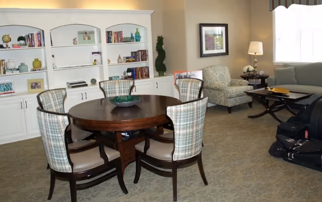 Communal living area with a round wooden table surrounded by four plaid-upholstered chairs, built-in white bookshelves, and upholstered seating by a window.