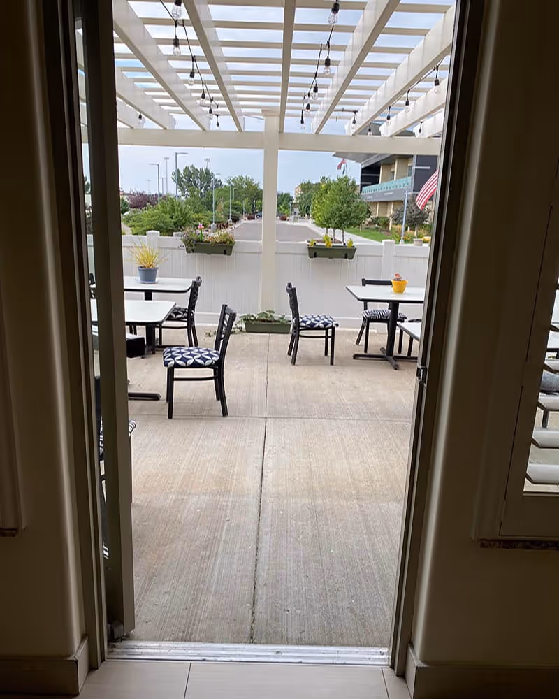 View through an open doorway leading to an outdoor patio area with tables and chairs. The patio has a white pergola overhead with string lights hanging. There are potted plants on the tables and along the white fence surrounding the patio. Trees and a building with an American flag are visible in the background.
