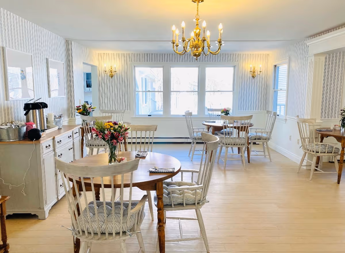 Bright communal dining room with round wooden tables, white spindle chairs, a chandelier, and a sideboard coffee station.