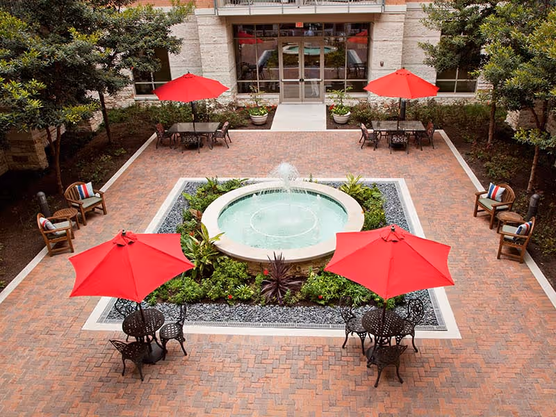 Outdoor courtyard area with a central circular water fountain surrounded by greenery. The courtyard is paved with brick and features several black metal tables with red umbrellas and matching chairs. There are also wooden chairs with cushions along the sides near trees and plants. The entrance to a building is visible in the background.