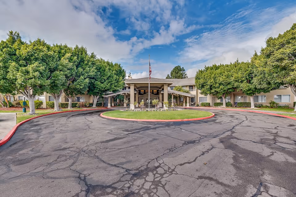 Front exterior view of Arcadia Place Senior Living facility showing a circular driveway with a flagpole in the center, surrounded by green trees and a beige two-story building under a partly cloudy sky.