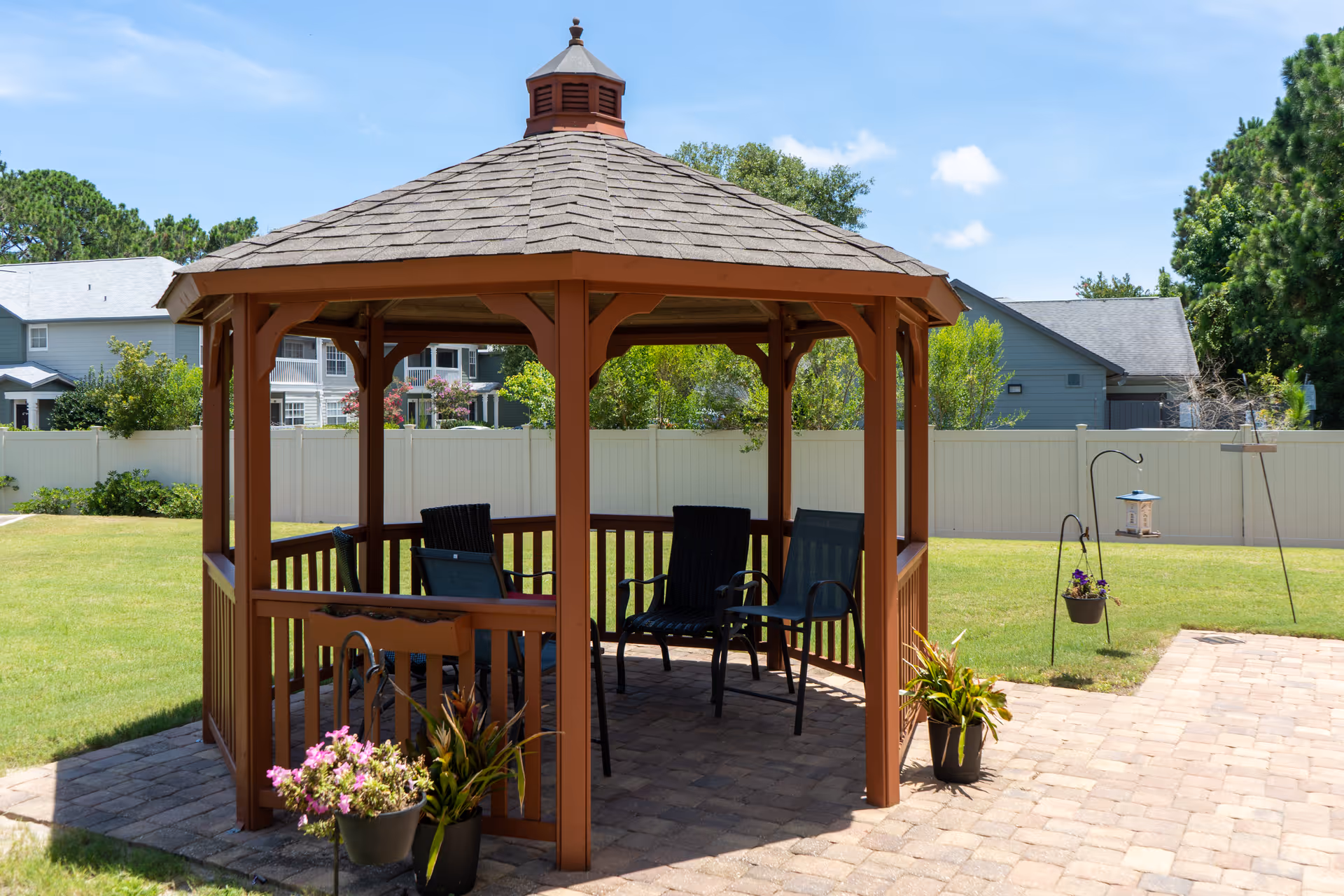 A wooden gazebo with chairs and potted plants on a paved patio in a grassy backyard with houses and a white fence behind it.