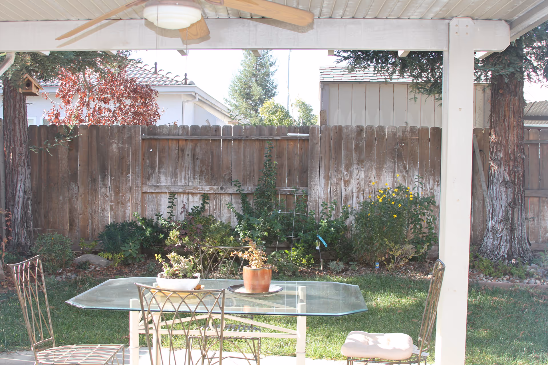 A covered outdoor patio area with a glass-top table and metal chairs, some with cushions. There are potted plants on the table and a wooden fence with greenery and trees in the background.