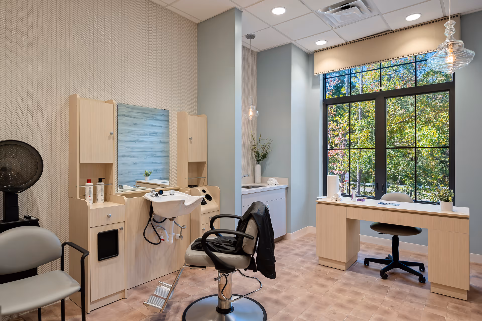 A bright and modern salon room with a hair washing station, salon chair, and a large window showing green trees outside. The room also features a desk with a rolling chair, a hair dryer, and light-colored wood cabinetry.