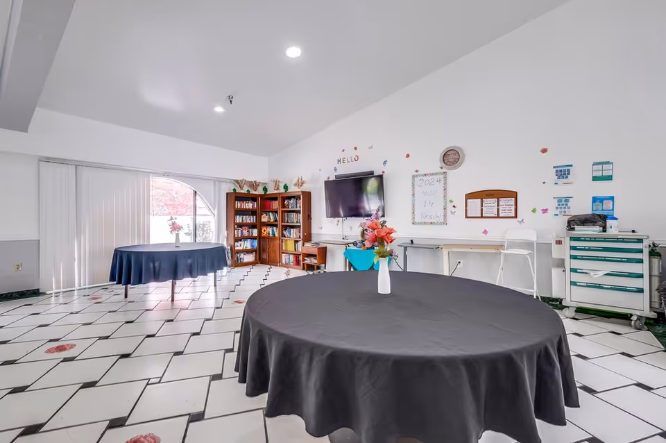 A bright room with two round tables covered with black tablecloths, each with a vase of flowers on top. The room has white walls decorated with colorful stickers and a wall-mounted TV. There is a wooden bookshelf filled with books near a large window with vertical blinds. The floor has a white tile pattern with black grout and red flower accents. A clock and various notices are displayed on the wall, along with a whiteboard showing the date May 14, 2024.