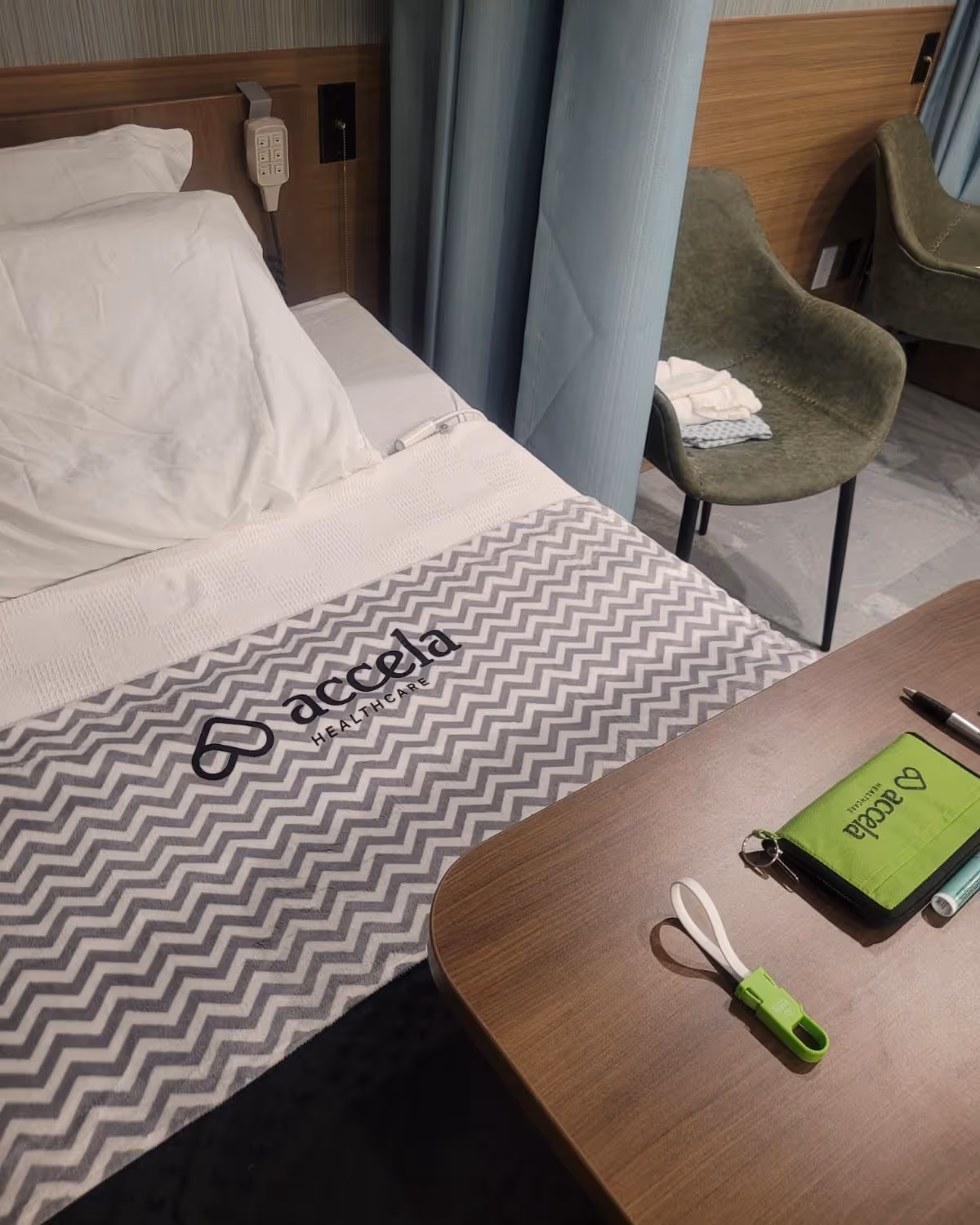 A neatly made bed with a white pillow and a chevron-patterned blanket featuring the Accela Healthcare logo. Next to the bed is a wooden table with a green Accela Healthcare pouch, a green USB drive, a pen, and a small tube. In the background, there is a green upholstered chair with folded towels on it, and a curtain partially drawn.