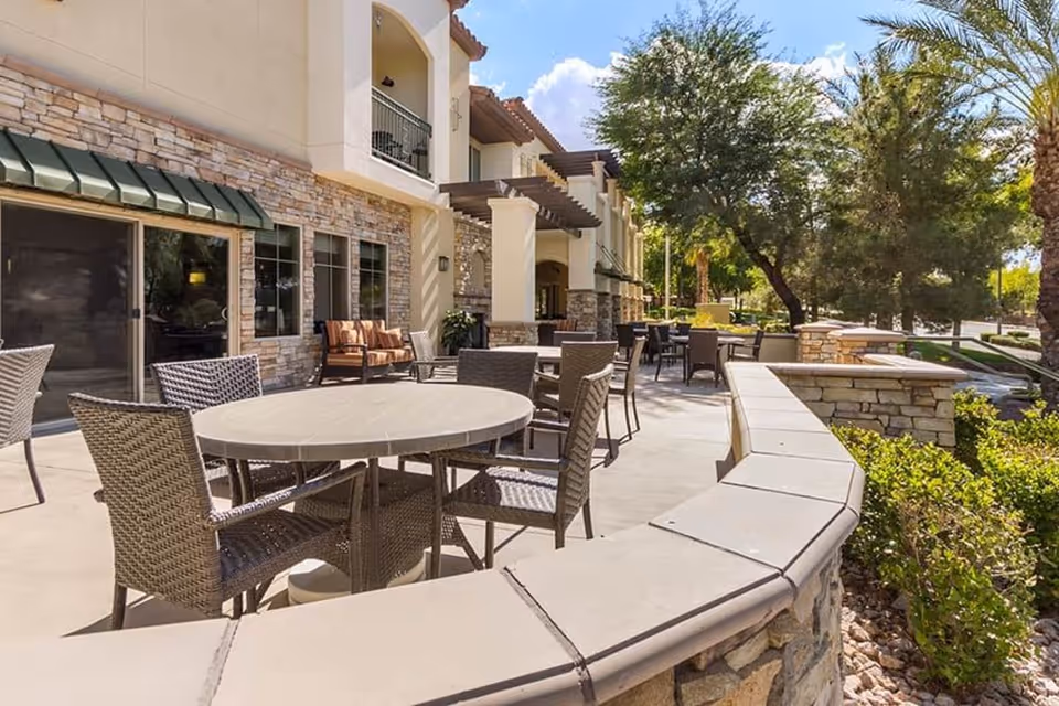 Outdoor patio with round tables and wicker chairs in front of a stone-faced senior living building surrounded by trees.