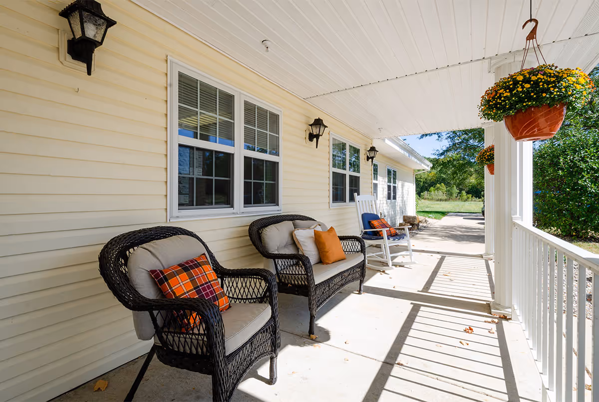 Covered front porch with wicker chairs, a rocking chair, cushions and hanging flower baskets.