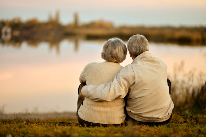 An elderly couple sitting on grass by a calm lake during sunset, with the man’s arm around the woman as they look out over the water.