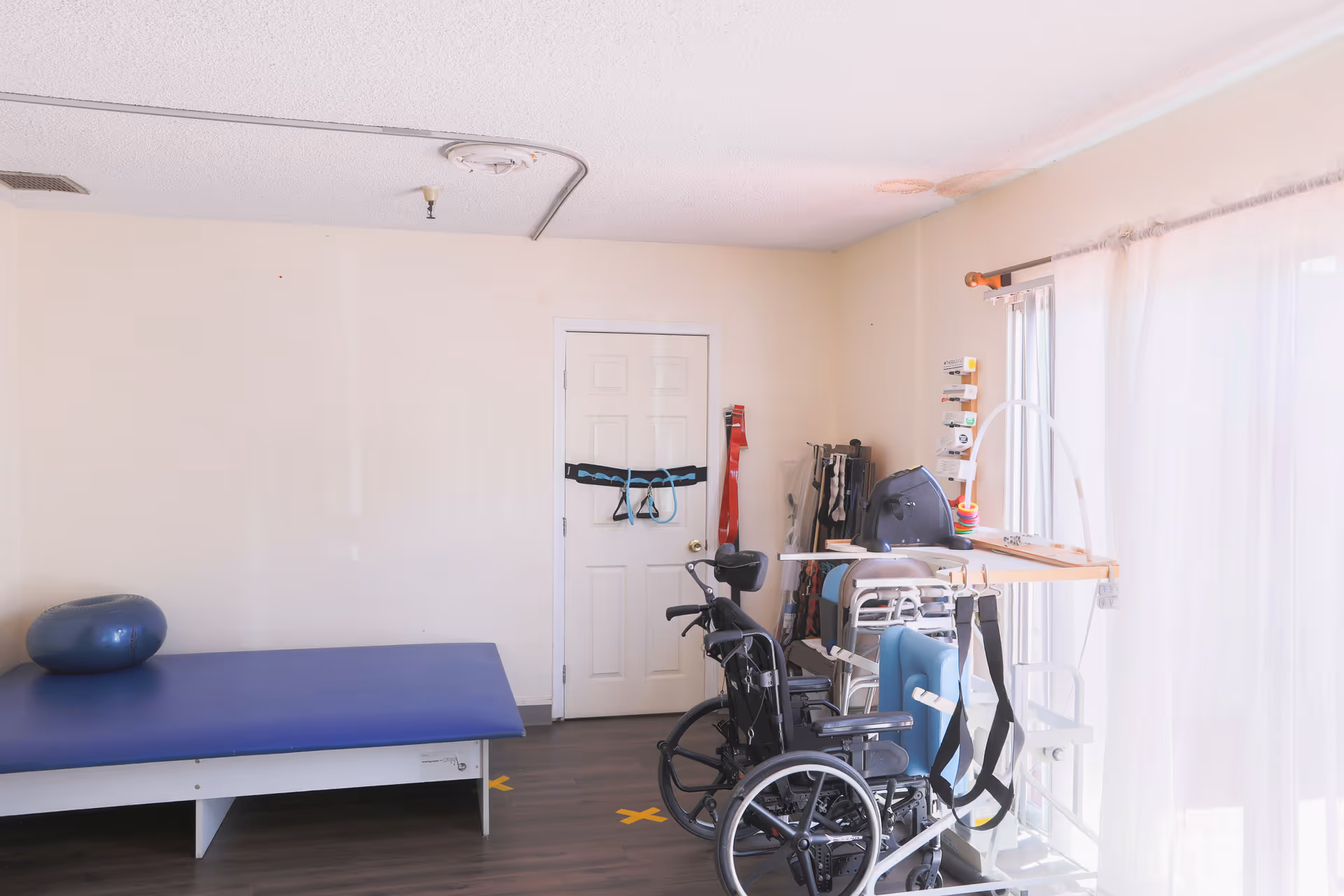 A small therapy or rehabilitation room with a blue padded therapy table, an exercise ball, a wheelchair, various mobility aids, and a door with exercise straps hanging on it. The room has light-colored walls, a wooden floor, and sheer curtains letting in natural light.