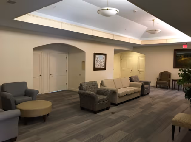 A spacious living room area with multiple armchairs and a beige sofa arranged along the walls. The room has a carpeted floor, neutral-colored walls, and ceiling lights. There are two archways and several closed doors visible, along with framed artwork on the walls.