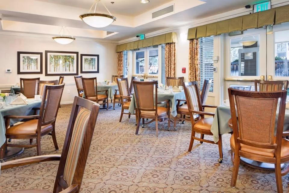 A well-lit dining room with multiple round tables covered with light green tablecloths, each set with napkins and cups. Wooden chairs with cushioned seats surround the tables. The room features patterned carpet, framed artwork on the walls, large windows with curtains, and ceiling light fixtures.