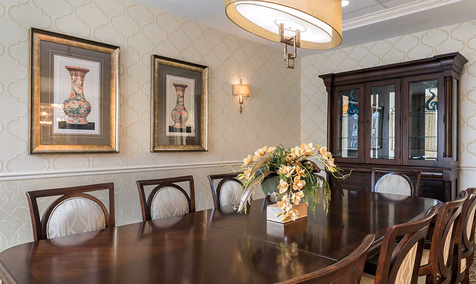 A formal dining room with a long polished wooden table surrounded by eight chairs with cushioned backs. A floral centerpiece decorates the middle of the table. On the wall, there are two framed pictures of decorative vases and a wall-mounted light fixture. A large wooden china cabinet with glass doors stands against the far wall. A modern circular chandelier hangs from the ceiling.