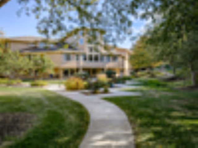 Curved walkway through a landscaped lawn leading to a two-story beige senior living building surrounded by trees.