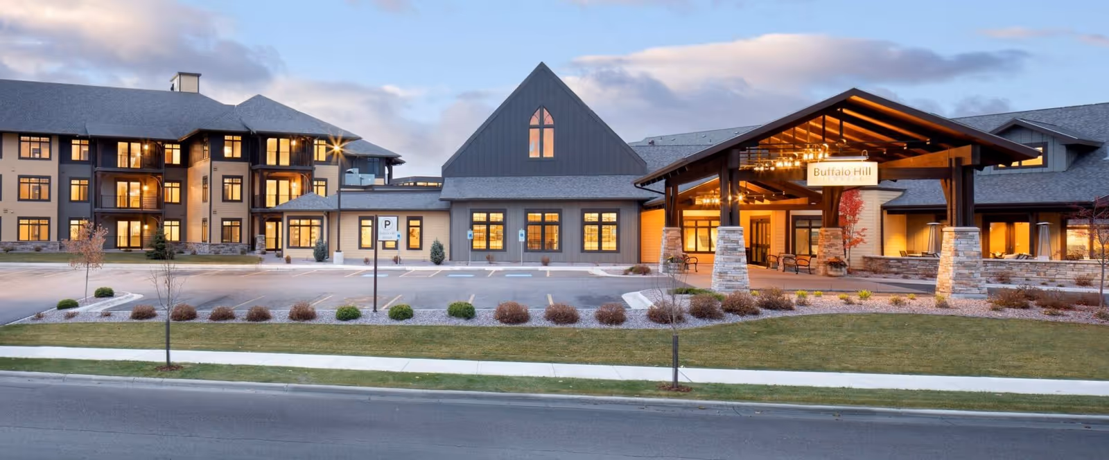 Exterior view of Immanuel Living at Buffalo Hill facility during dusk, showing a large building with multiple windows lit from inside, a covered entrance with stone pillars, and a parking area in front.