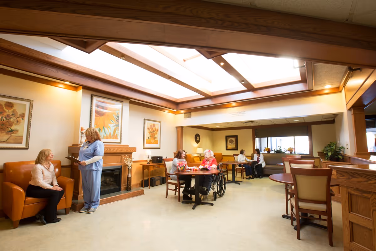 A bright communal lounge in a senior living facility with residents and staff seated around tables near a fireplace under skylights.