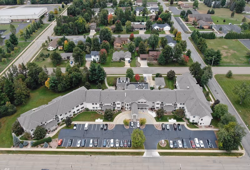 Aerial view of Renaissance by Rennes senior living facility in Wisconsin Rapids, showing a large, U-shaped building with a gray roof surrounded by trees and parking lots. Residential houses and streets are visible in the background.