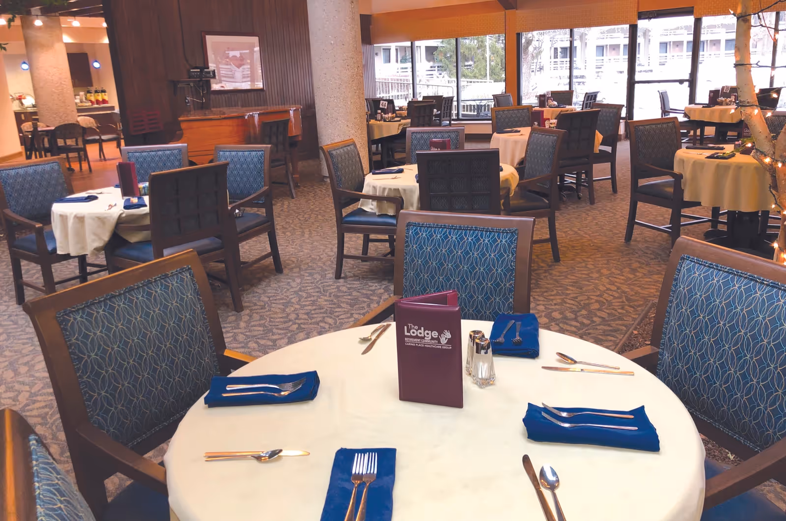 Dining room at The Lodge Retirement Community with round tables set with blue napkins and place settings and upholstered chairs.