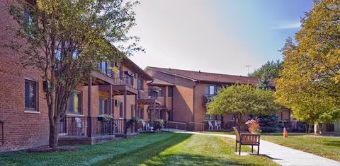 Exterior view of a senior living facility with brick buildings, balconies, trees, a grassy lawn, a sidewalk, and a bench under a clear blue sky.