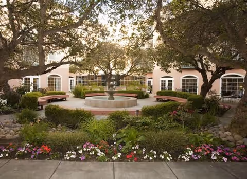 A peaceful outdoor courtyard with a central water fountain surrounded by benches, trees, and colorful flower beds, with a light pink building featuring multiple windows in the background.