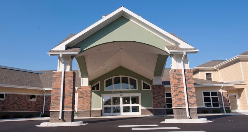 Front exterior view of a senior living facility entrance with a covered drop-off area supported by brick and white columns, green and beige walls, and a clear blue sky above.