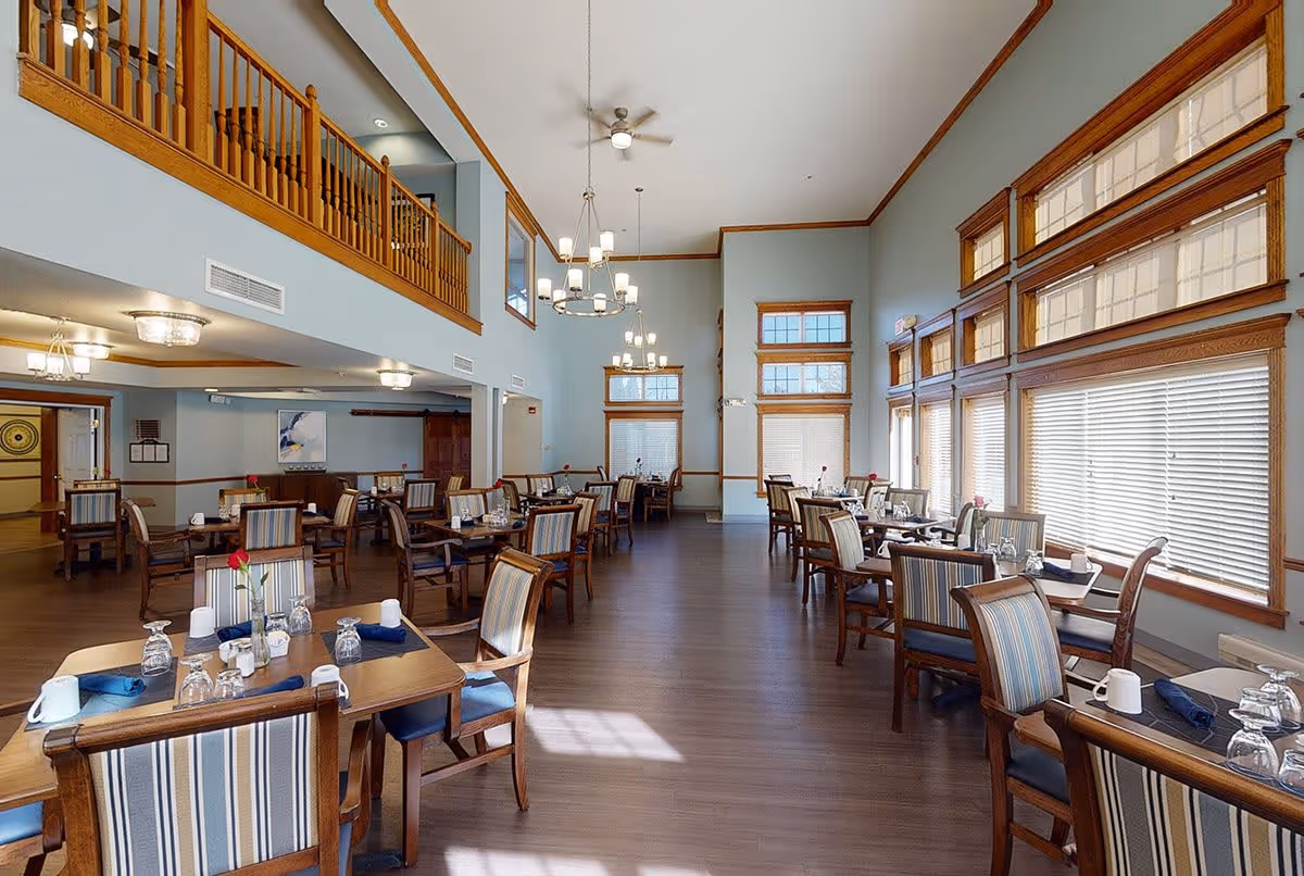 A spacious dining room with multiple wooden tables and chairs arranged neatly. Each table is set with glasses, napkins, and a single red rose in a vase. The room features large windows with blinds, light blue walls with wooden trim, and high ceilings with ceiling fans and chandeliers. There is a wooden balcony railing visible on the upper level.