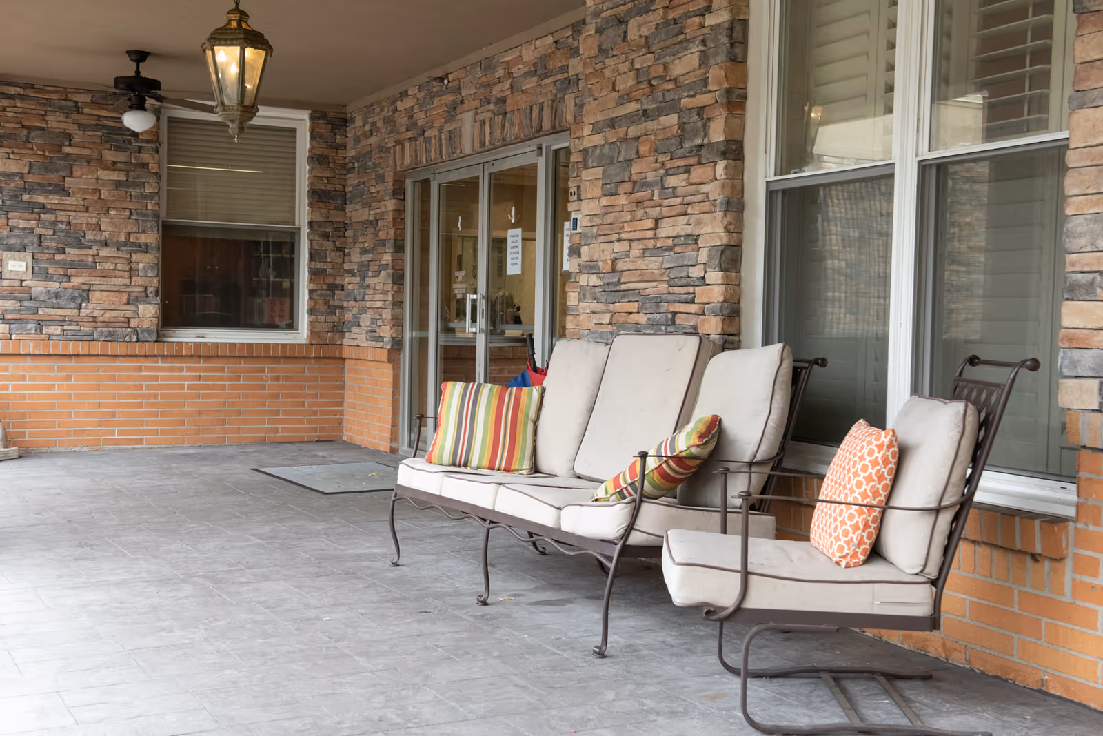 Outdoor covered patio area with cushioned metal seating including a three-seat bench and a single chair, both with decorative pillows. The area has stone and brick walls, a hanging lantern light, a ceiling fan, and large windows and glass doors leading inside.