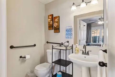 Small accessible bathroom with a pedestal sink, toilet, grab bars, mirror, light fixture and a black metal shelving unit; a bedroom is visible through the open door.