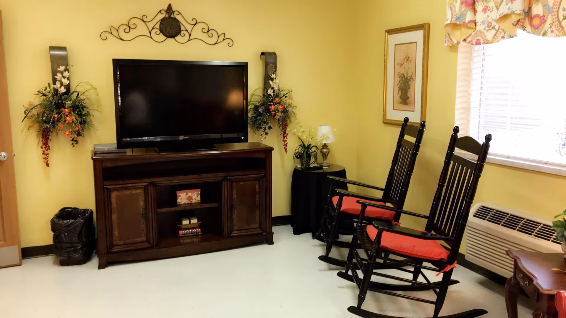 A cozy room with yellow walls featuring a flat-screen TV on a wooden cabinet, two black rocking chairs with red cushions, floral wall decorations, a small round table with a lamp and plant, a framed picture on the wall, and a window with floral curtains letting in natural light.