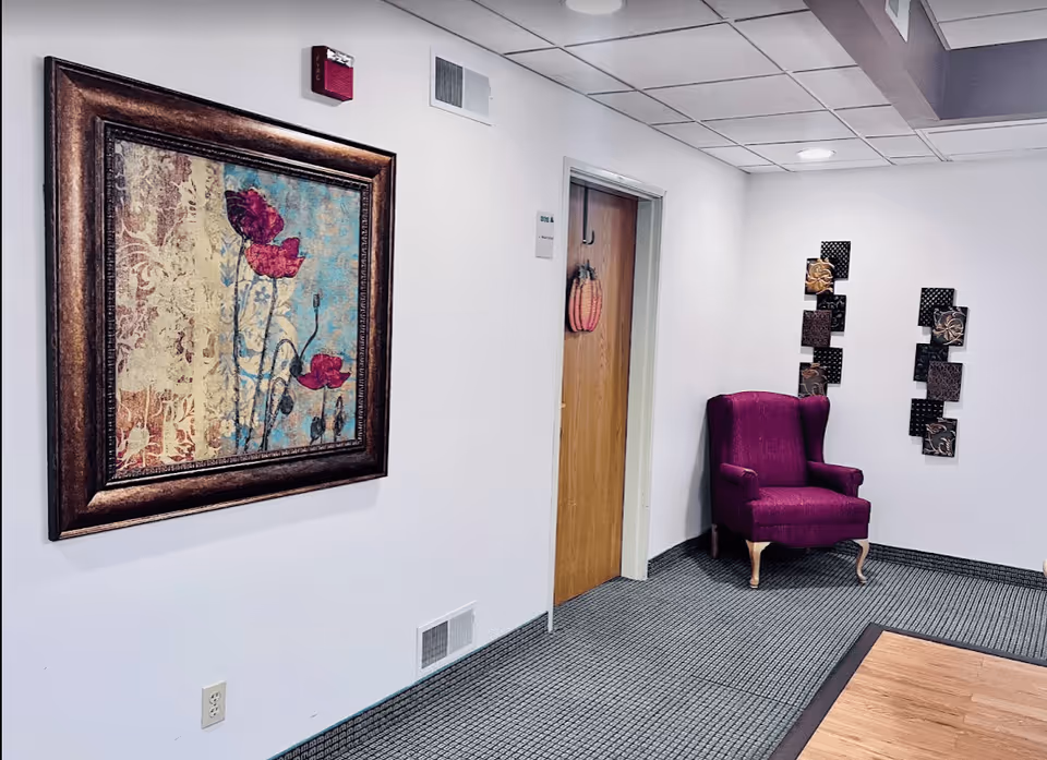 Interior hallway of a senior living facility with a framed floral painting on the left wall, a wooden door decorated with a pumpkin hanging, a purple upholstered armchair in the corner, and decorative wall art on the right wall. The floor is carpeted with a patterned design and the ceiling has recessed lighting.