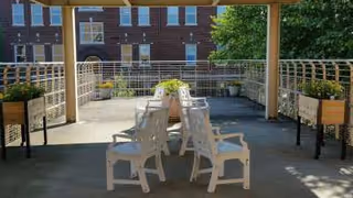 Outdoor patio area with white chairs arranged around a rectangular table under a pergola. Raised garden beds with plants are on either side, and a brick building is visible in the background.