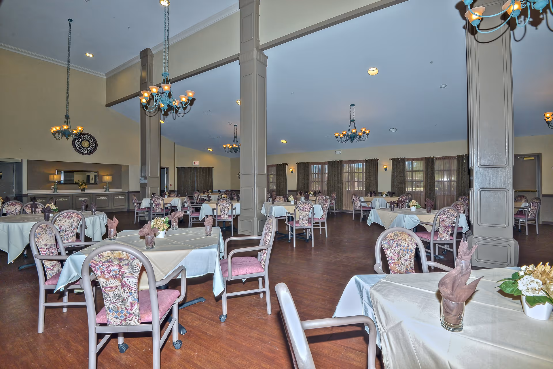 A spacious dining room with multiple tables covered in white tablecloths, each set with folded napkins and small flower arrangements. The room features wooden floors, floral upholstered chairs, large windows with curtains, and several chandeliers hanging from the ceiling.