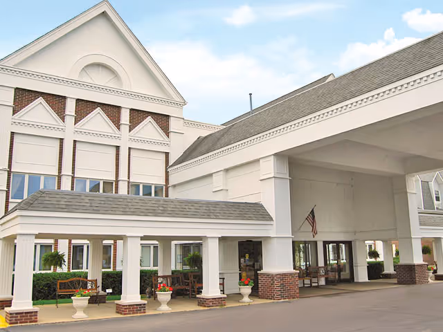 Front entrance of a Victorian-style building with a covered porte-cochere, columns, benches, planters, and an American flag.