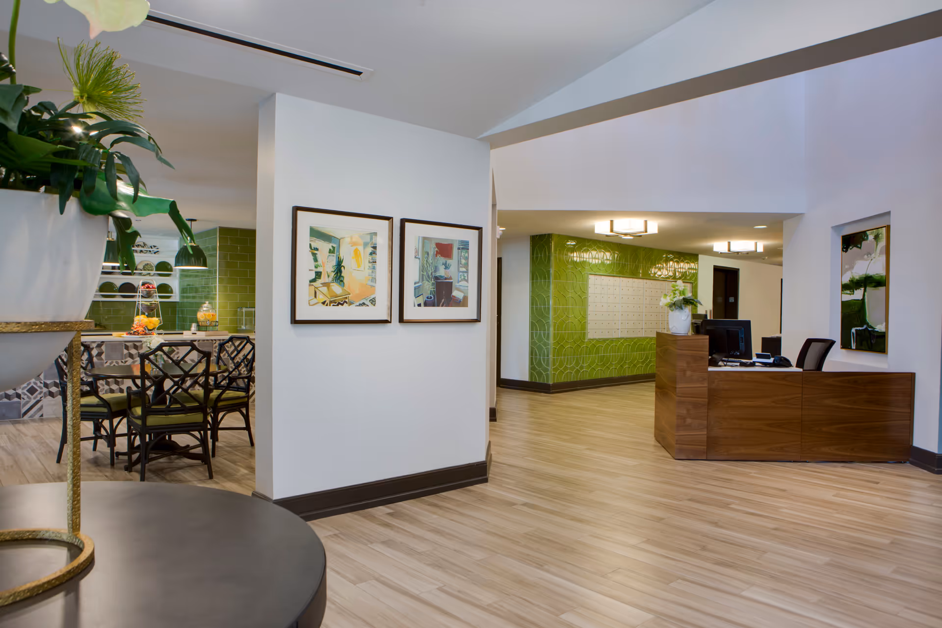 Interior view of a senior living facility lobby area with a wooden reception desk on the right, green tiled wall with mailboxes in the background, and a dining area with a table and chairs on the left. The space features light wood flooring, white walls, framed artwork, and modern ceiling lights.