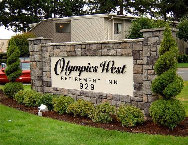 Stone sign for Olympics West Retirement Inn with the address number 929, surrounded by neatly trimmed bushes and spiral-shaped topiary trees, with part of a building and parked cars visible in the background.