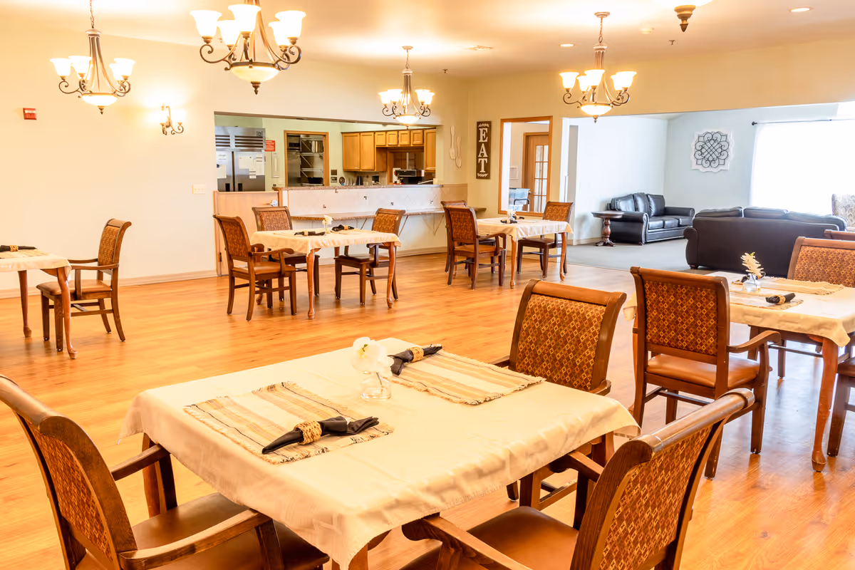 A bright dining room with wooden floors and several tables covered with beige tablecloths, each set with napkins and small flower vases. The room features multiple chandeliers hanging from the ceiling. In the background, there is an open kitchen area with wooden cabinets and stainless steel appliances, and a seating area with dark leather sofas near a large window.
