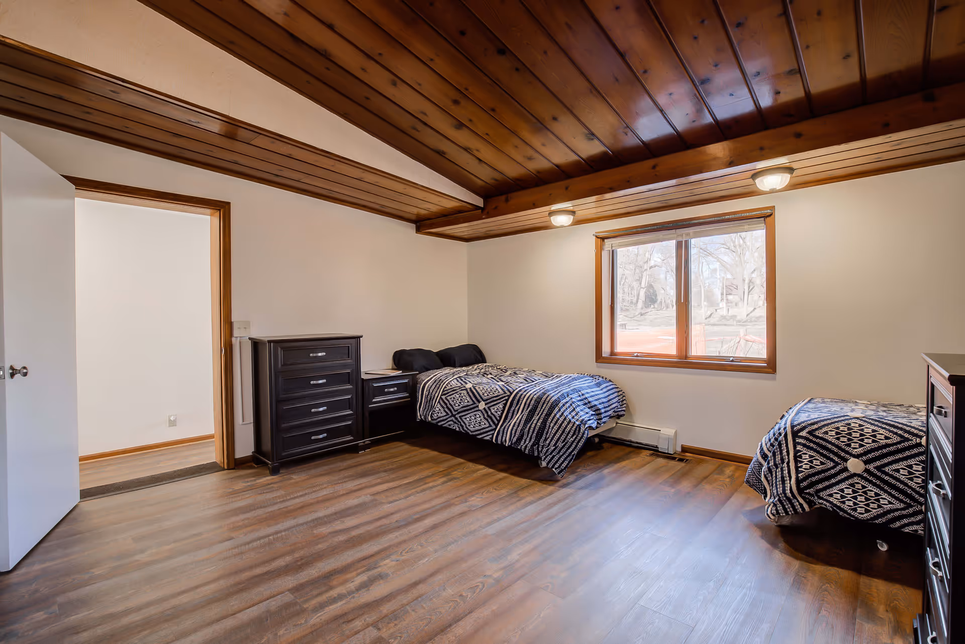 A bedroom with two single beds covered in black and white patterned bedding, a wooden ceiling with recessed lighting, a large window with a view of trees outside, and dark wood furniture including a chest of drawers and a nightstand.