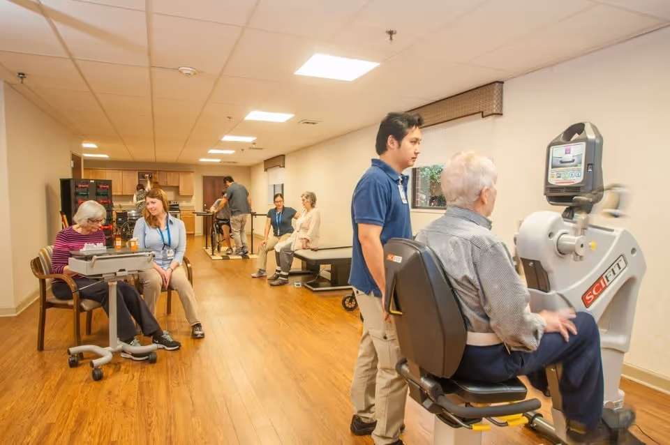 A rehabilitation room with elderly residents and staff members. One elderly man is seated on a SciFit exercise machine being assisted by a staff member. In the background, other residents are engaged in physical therapy activities, including walking with support bars and sitting on benches. The room has wooden flooring, beige walls, and bright overhead lighting.
