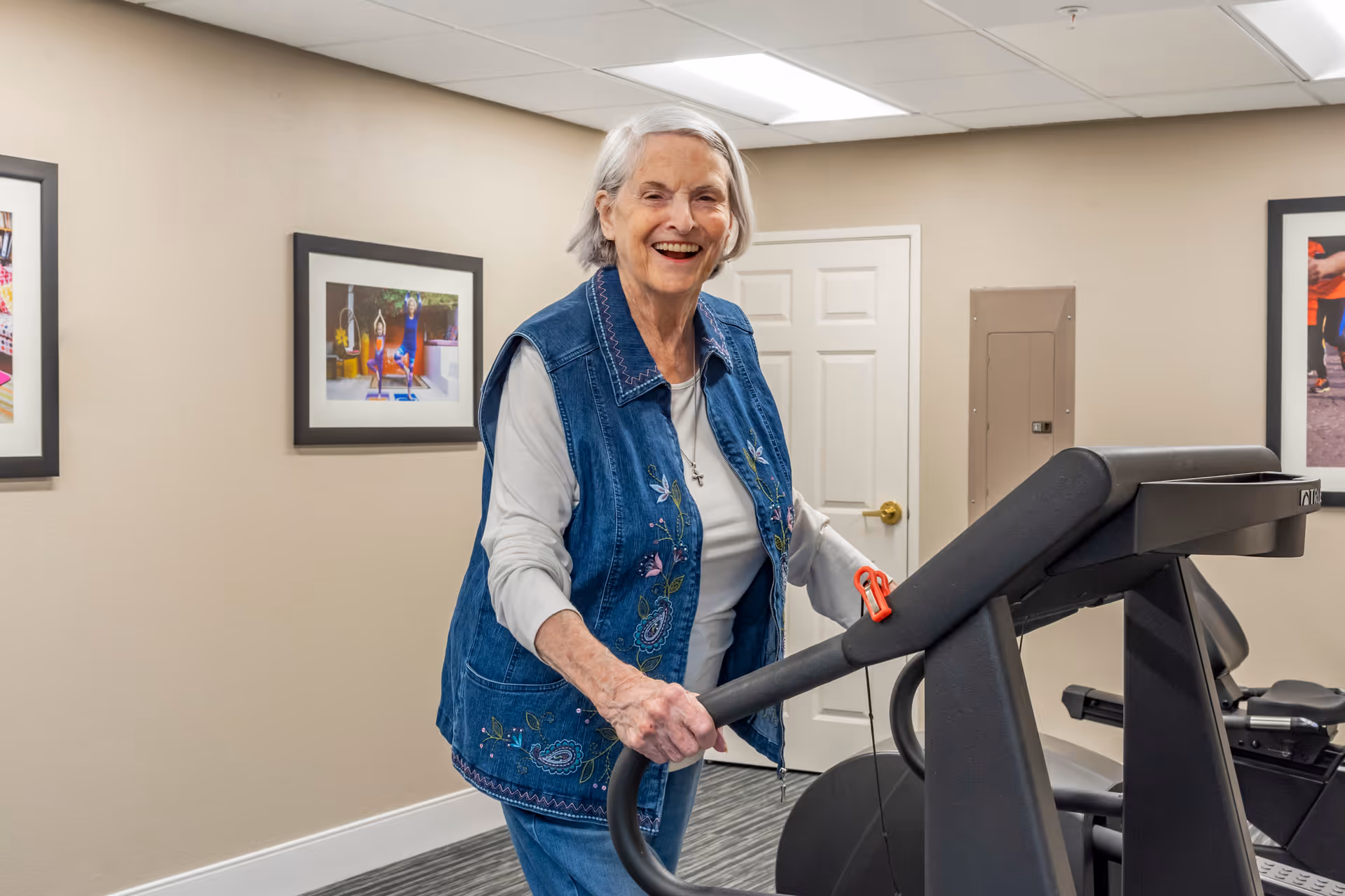 An elderly woman with gray hair wearing a blue embroidered vest and white shirt is smiling while standing on a treadmill in a fitness room. The room has beige walls with framed pictures and a white door in the background.