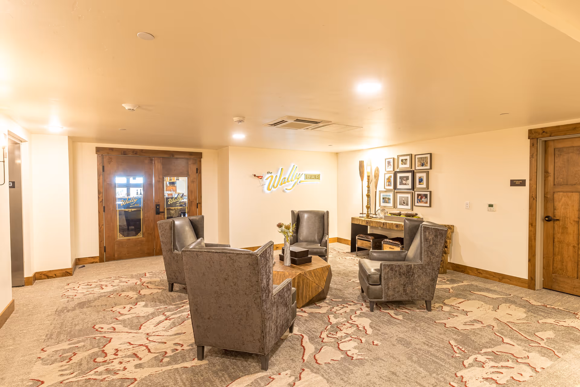 A cozy seating area with four gray armchairs arranged around a wooden coffee table on a patterned carpet. The room has warm lighting and wooden trim. On the wall, there is a neon sign that reads 'the Wally Bar' and a console table with decorative items and framed photos above it. Double wooden doors with glass panels are visible in the background.